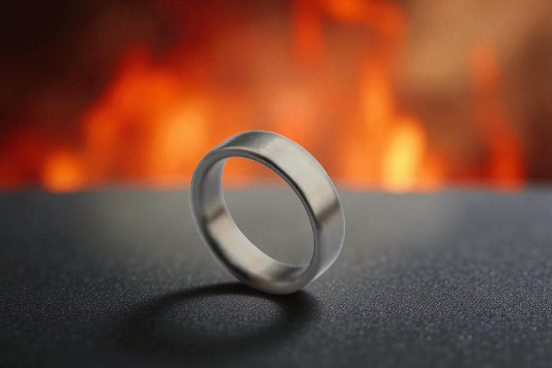 Close-up of brushed titanium flat-edge ring on dark textured surface with firelight reflection