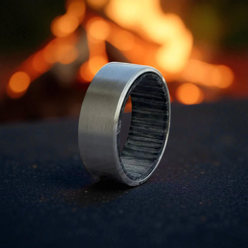 Titanium ring with brushed matte finish and grey whiskey barrel wood interior, displayed against dark surface with firelight background