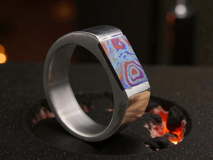 Titanium D-shaped ring featuring vibrant zirconium inlay with layered blue and purple patterns, placed on molten charcoal surface