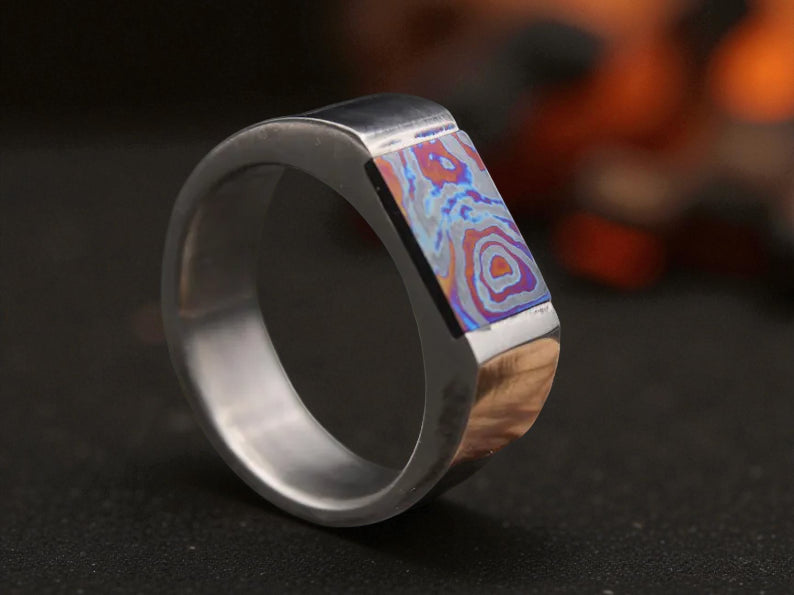 Close-up of geometric titanium D ring with zirconium Damascus-style inlay reflecting firelight background