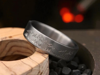 Titanium ring with deep engraved mountain and forest landscape placed on wooden accent beside dark stones