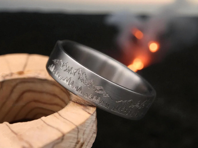 Close-up of matte titanium band with engraved mountain and pine tree motif, glowing fire background