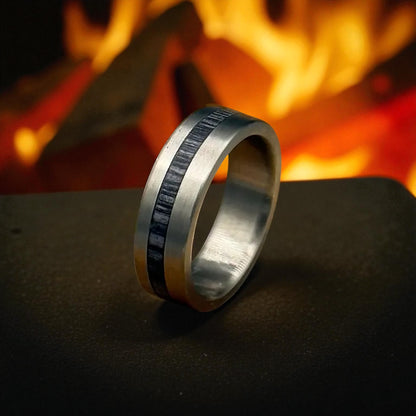 Titanium ring with brushed silver exterior and grey whiskey barrel wood inlay, positioned on a dark matte surface with a fire-lit background