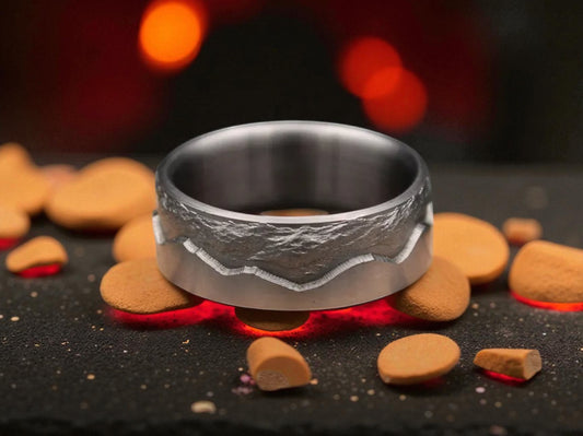 Titanium ring with carved mountain peak design on the upper edge, placed on dark volcanic stones with warm firelight in the background